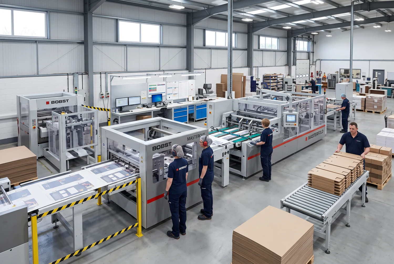 Công nhân đứng cạnh máy gia công carton và các chồng tấm bìa phẳng trong nhà máy